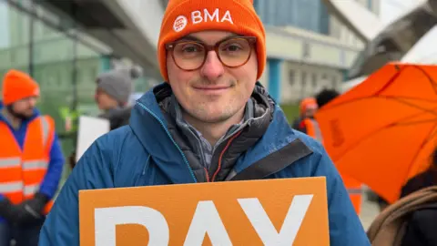 A man with an orange beanie on that has white writing that reads "BMA". He is wearing a blue waterproof jacket and is standing outside a glass hospital building