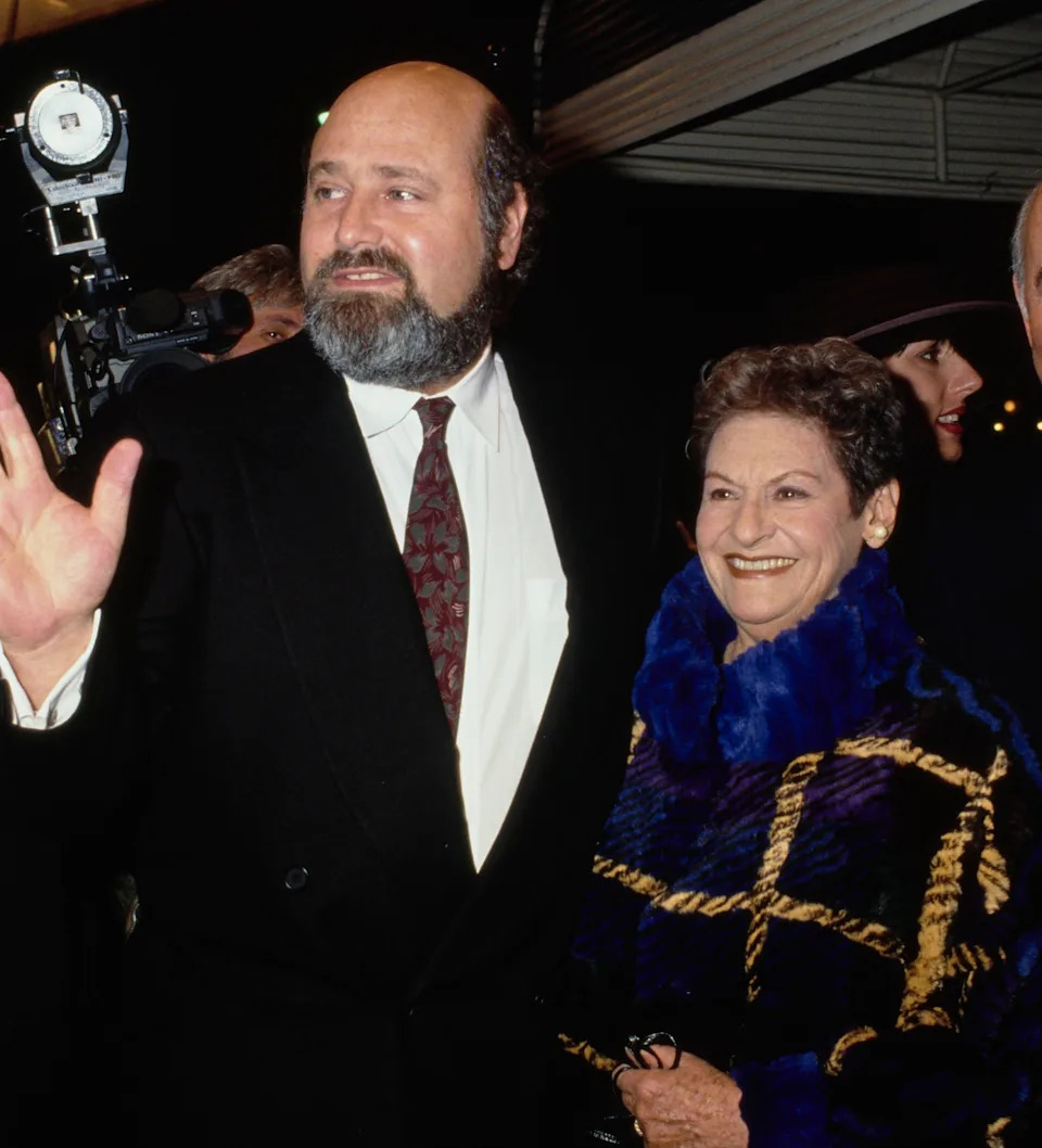 Four people pose on a red carpet; one waves. They wear formal attire, including suits and a patterned coat. They appear pleased