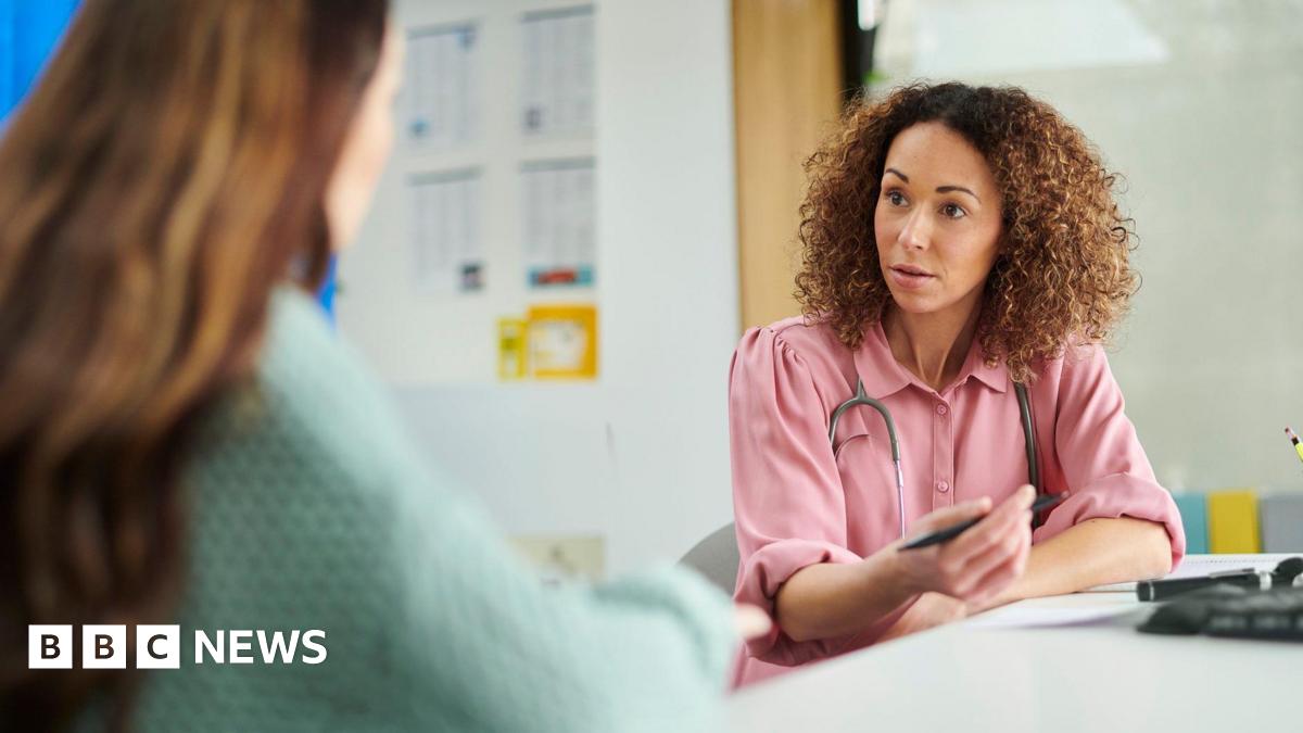 A female GP wearing a pink blouse and a stethoscope around her neck, talks to a female patient with long, brown hair (who is blurred and the back of her head is visible only in the foreground)