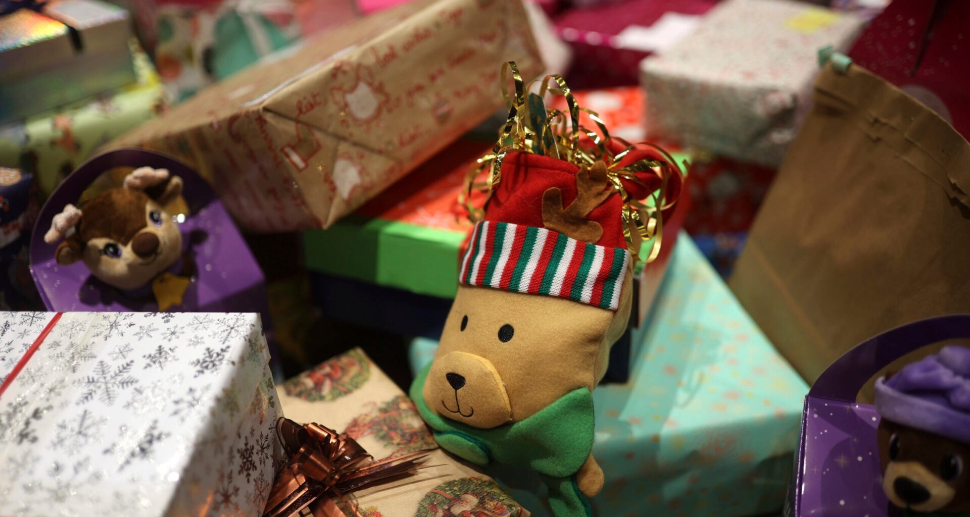 A variety of wrapped gifts and boxes, including a plush bear toy wearing a striped hat, are arranged in a festive setting.