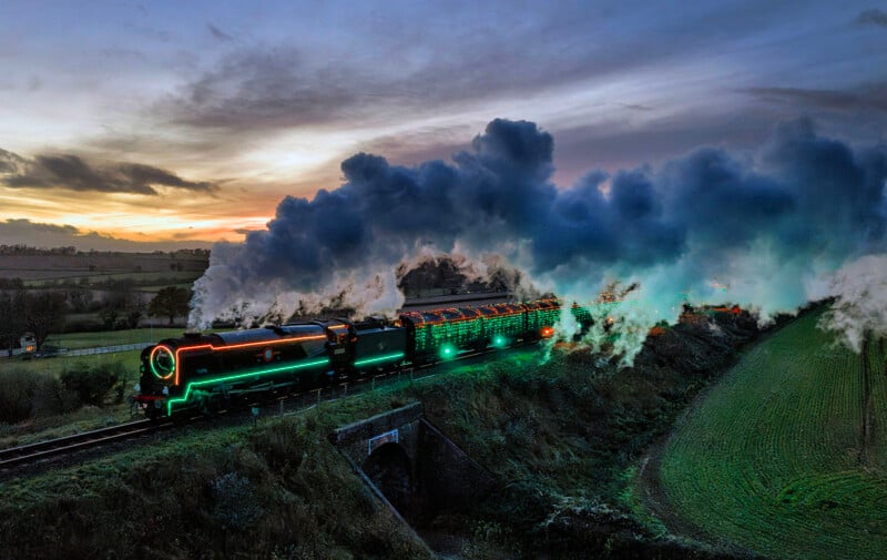 A steam train decorated with colorful Christmas lights travels through the countryside at dusk, billowing thick white smoke under a dramatic, cloudy sky.