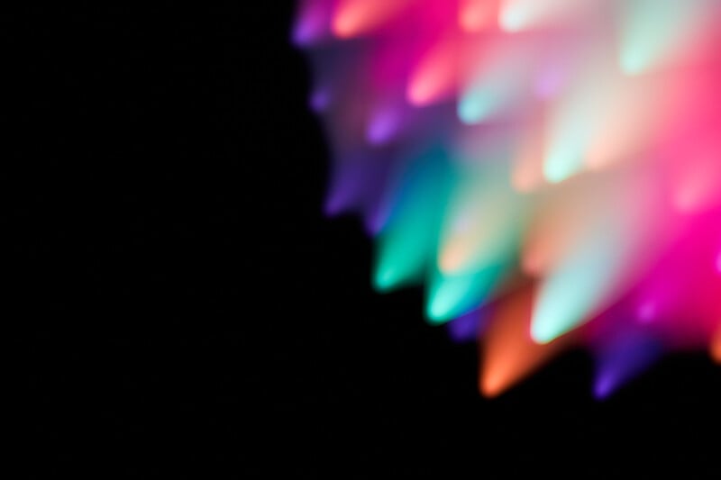 Abstract image showing blurred, colorful lights in shades of pink, purple, blue, green, and orange on a black background, creating a vibrant, glowing pattern on the right side of the frame.