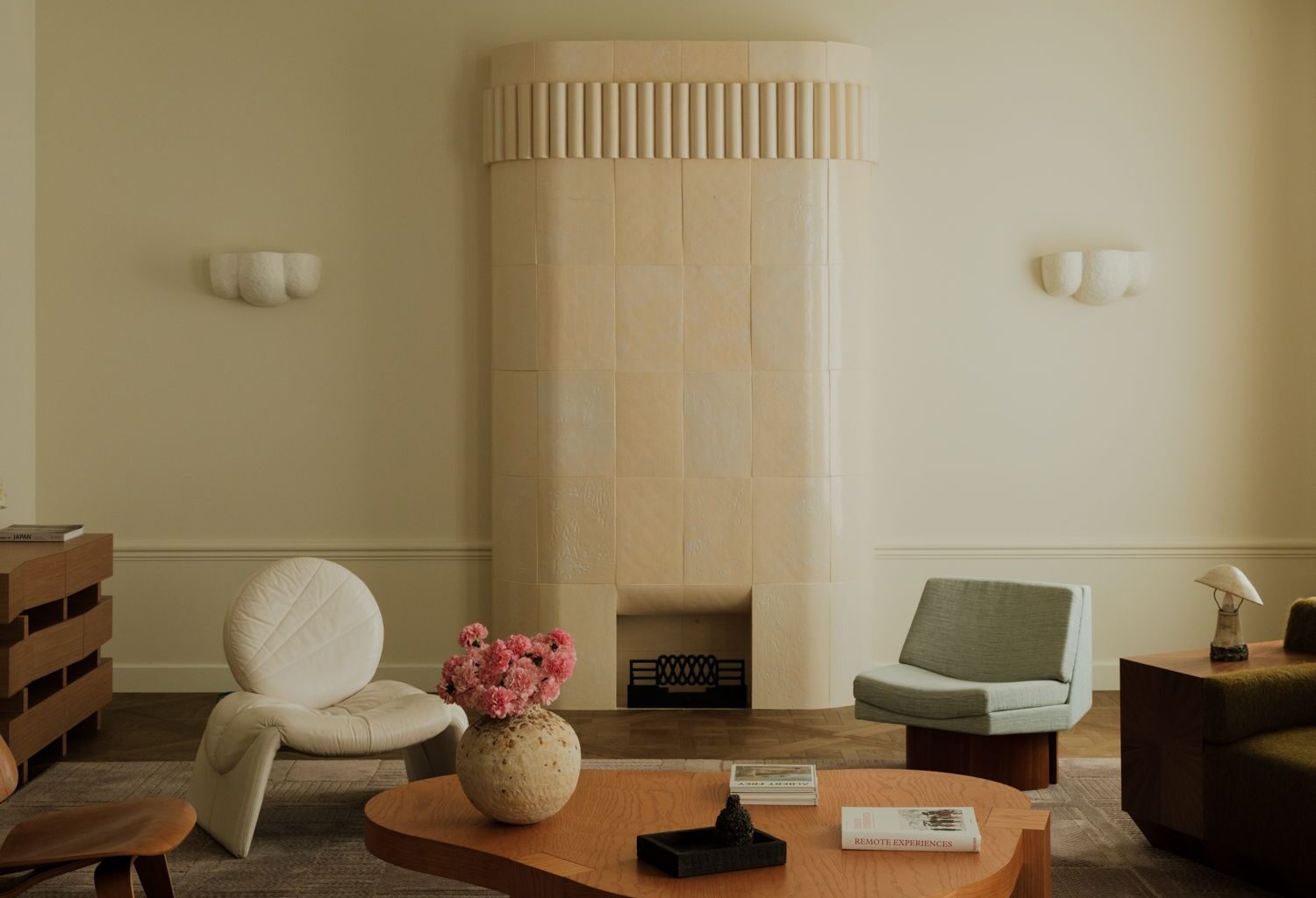 Image of a soft minimalist living room with white walls and curved shapes. There is a pastel stone, rounded fireplace, a curved wooden coffee table, and a mix of accent chairs around the room.