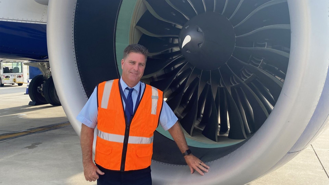A man in an orange safety vest stands confidently beside a large aircraft jet engine on an airport tarmac, conveying a sense of professionalism. He uses V A's telehealth services.