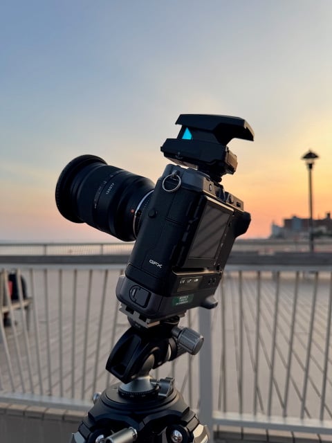 A professional camera mounted on a tripod is set up outdoors near a railing, capturing a scenic sunset with soft pastel skies and a streetlamp in the background.