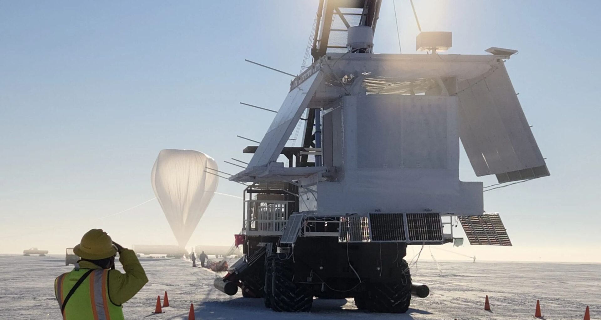 An image from Antarctica shows a giant machine in the foreground with a construction worker with a vest in the front left image and a white weather balloon in the background