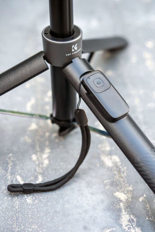 Close-up of a black tripod with a remote control attached to one of its legs, positioned on a textured gray surface. The brand "K&F Concept" is visible on the central part of the tripod.