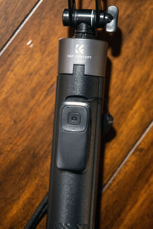 Close-up of a black K&F Concept tripod or selfie stick handle with a built-in camera remote button, resting on a wooden surface.