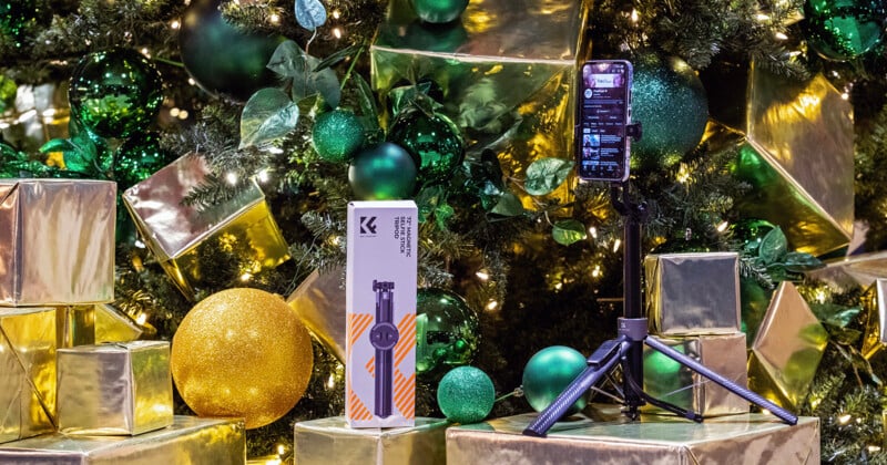 A smartphone mounted on a tripod stands next to its packaging in front of a decorated Christmas tree with green and gold ornaments and wrapped presents.