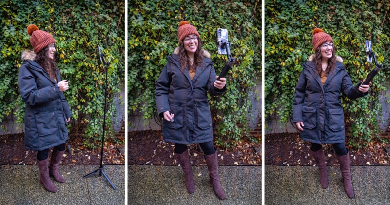 A woman in a winter coat, boots, and orange knit hat stands outdoors. She sets up a tripod with a phone, then takes a selfie, smiling, with green ivy covering the wall behind her.