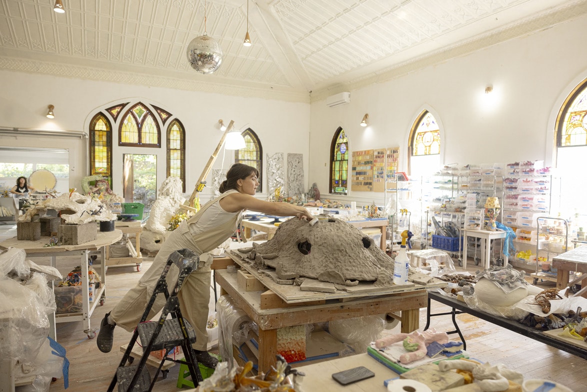 Artist working on a large sculpture in a bright, spacious studio with stained glass windows and various art materials.