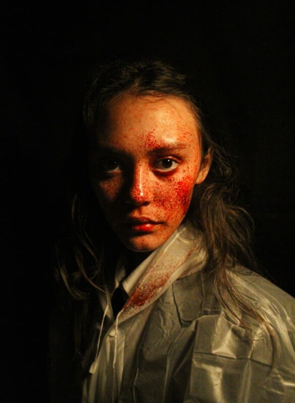 A person with long hair and a serious expression, wearing a white plastic gown, stands against a dark background. Their face is splattered with red spots resembling blood.