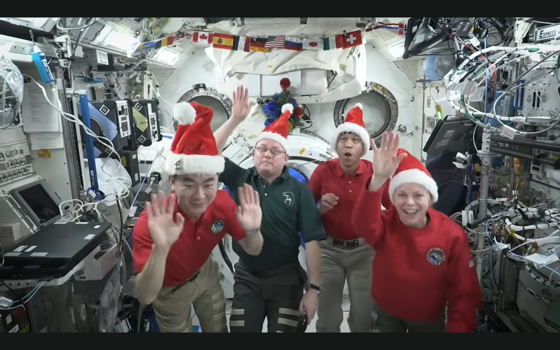 Four astronauts in Santa hats iwaving inside a space station decorated for Christmas