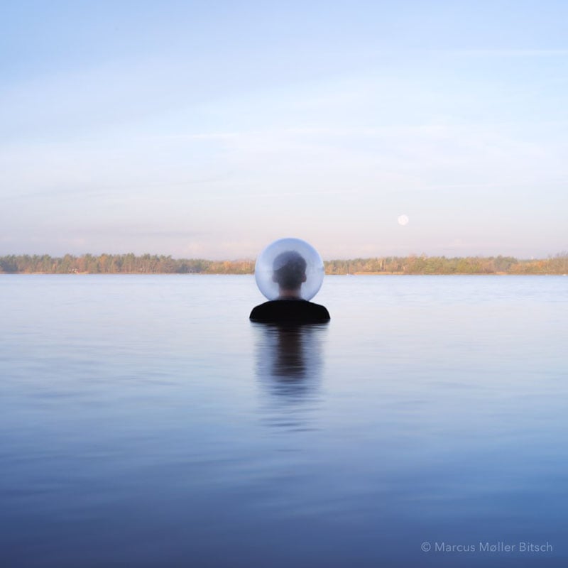 A person stands waist-deep in calm water, facing away, wearing a large transparent bubble helmet. The sky is clear, and distant trees line the horizon under soft daylight.