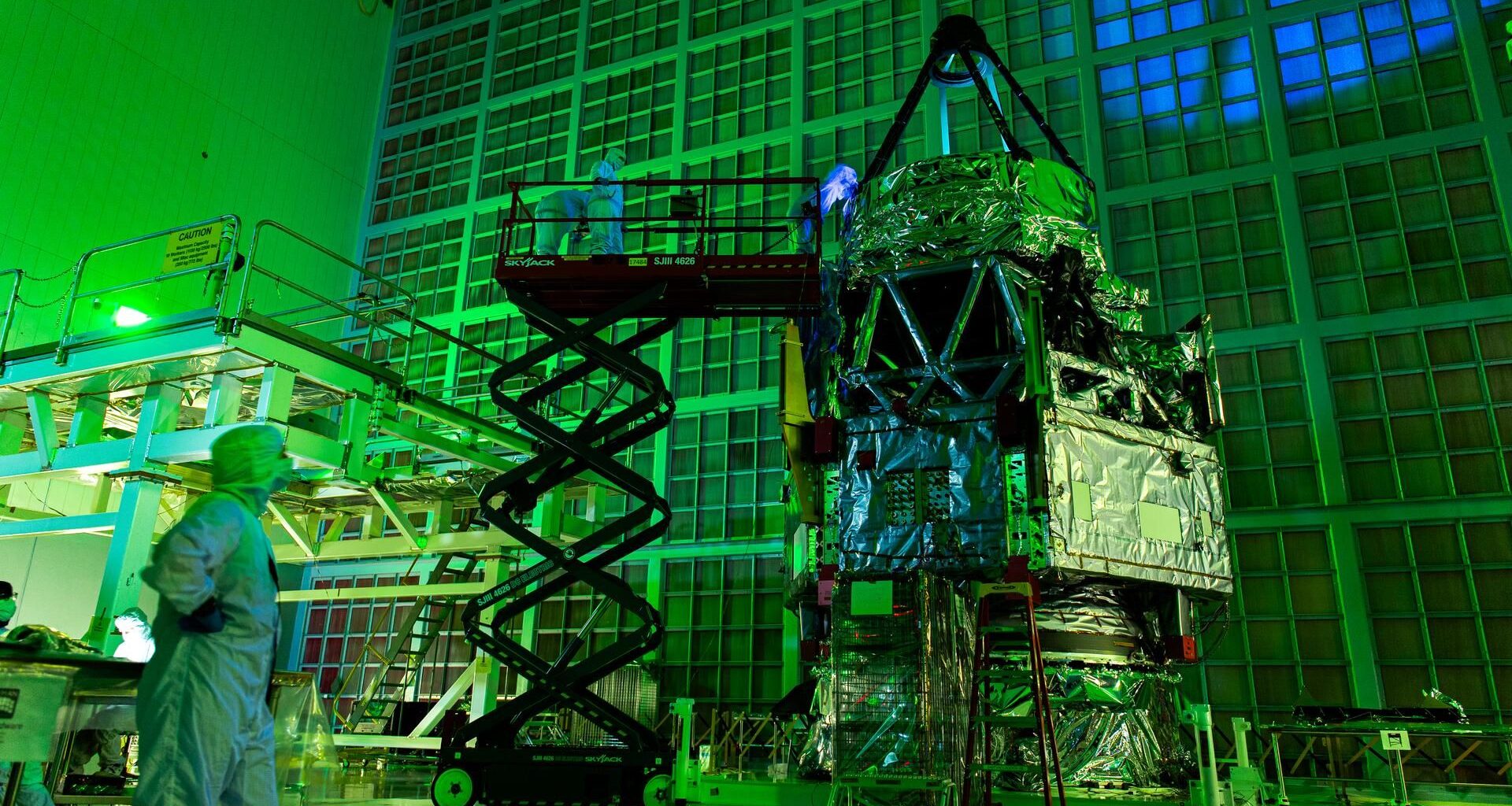 A telescope with a triangular top wrapped in foil stands next to a scaffold with people wearing white clean suits examining it under green light.