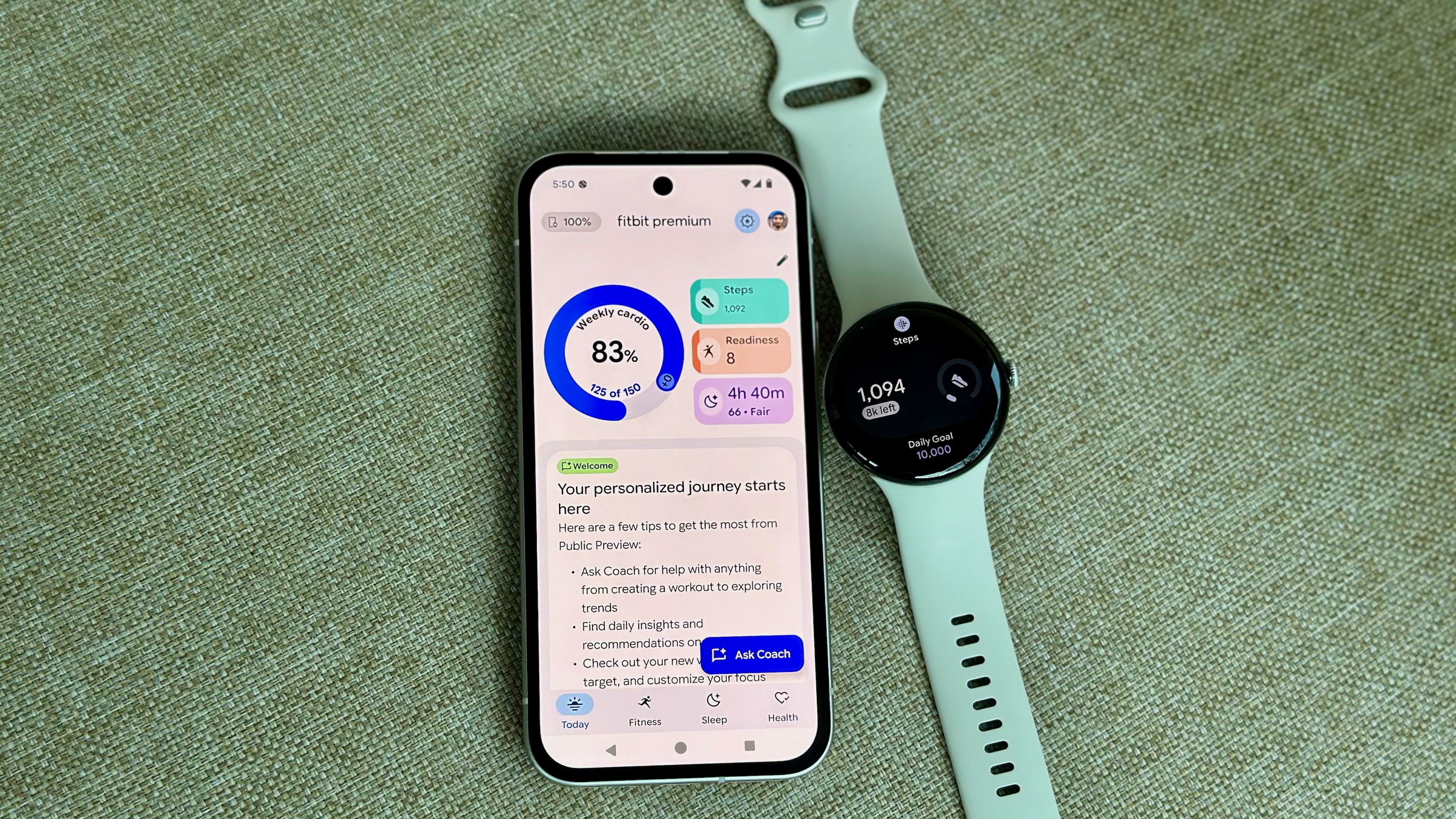 A photo of the Google Pixel Watch 4 sitting next to a Pixel 9a open to the Fitbit app, showing the Fitbit Premium Health Coach preview with the Today tab open; it shows new widgets and insights.