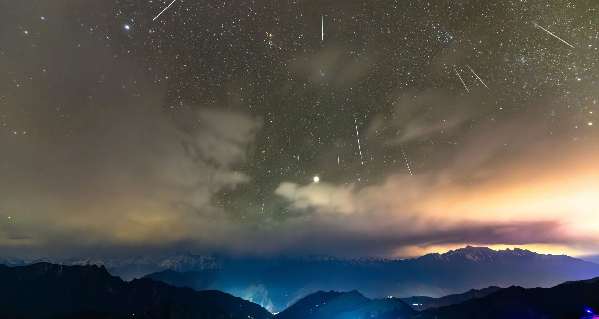 Shooting stars are pictured streaking downwards through a starry sky towards a cloudy horizon lined with mountains, lit in a blue mist.