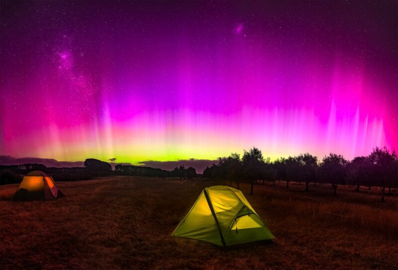 Stars seeping through magenta and yellow aurora with a tent lit from within in the foreground.