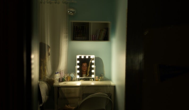A person sits at a desk in a dimly lit room, looking into a lighted mirror. Their reflection is visible. The desk has various items on it, and shelves with books are mounted on the wall above.