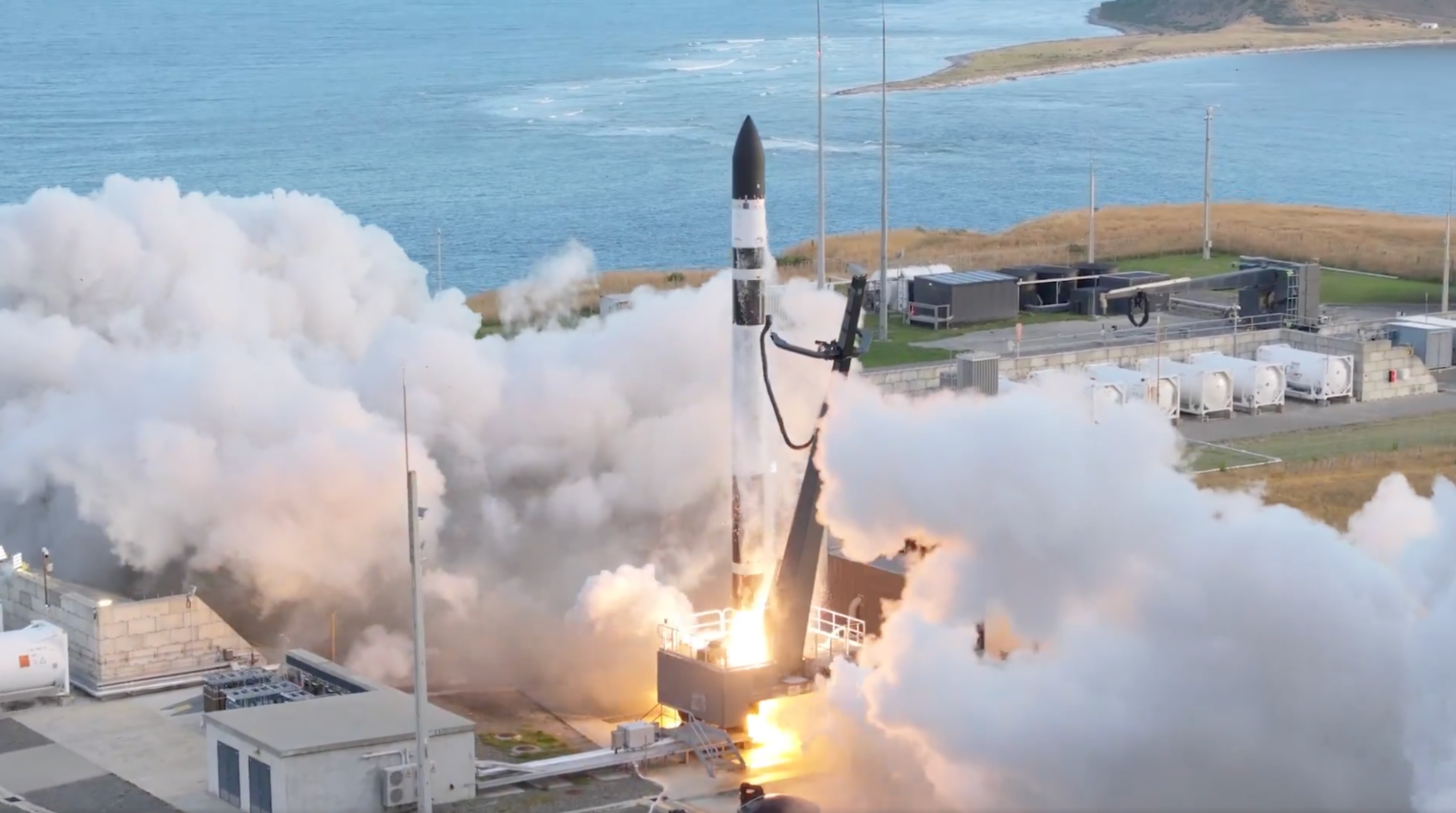 a black and white rocket launches from a seaside pad into blue skies