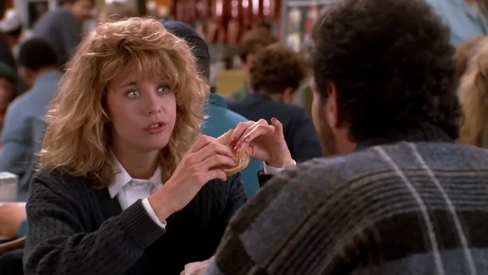 Woman with curly hair sits at a diner table holding a sandwich, talking to a man in a sweater, surrounded by other diners