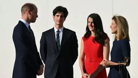 AFP via Getty Images Prince William is standing and chatting with Jack, Caroline and Tatiana Schlossberg. They are all wearing very dark suits and Tatiana is wearing a bright red dress.