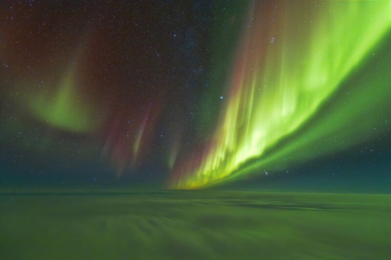 Clouds below, green and red curtains of aurora above with some stars shining through.