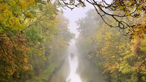 Josie Weller A narrowboat disappears into the distance on a misty day, with autumnal trees both sides of the canal 