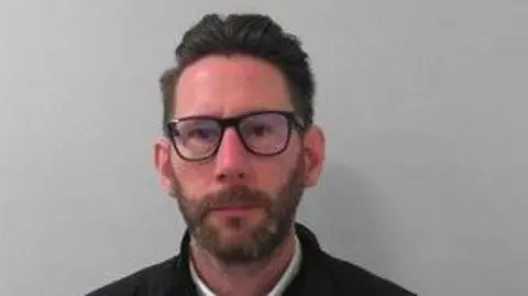 North Yorkshire Police A custody picture of a man with short dark hair, a beard, and glasses, wearing a white collared shirt under a dark sweater and a black quilted jacket, standing against a plain light grey wall.