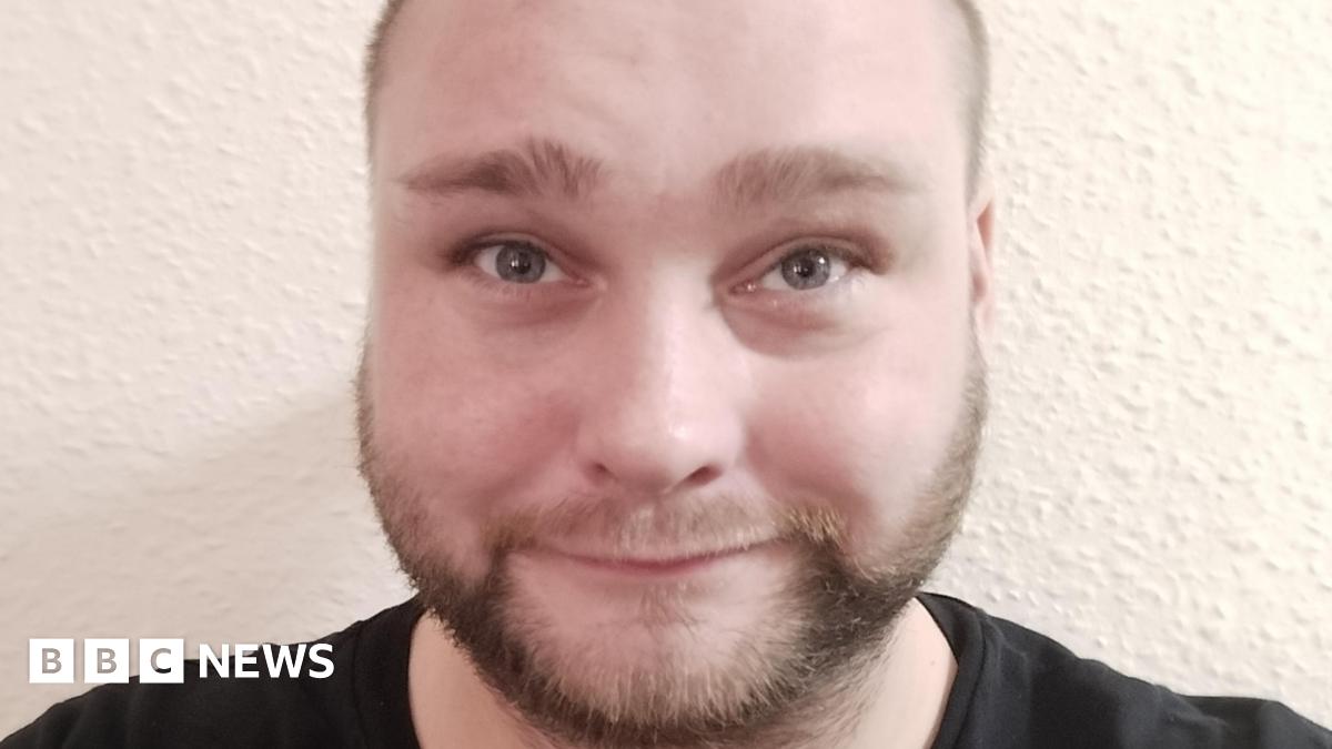 A close-up selfie of a man with a light brown beard and light-brown eyes. He is smiling at the camera. There is a white wall behind him.