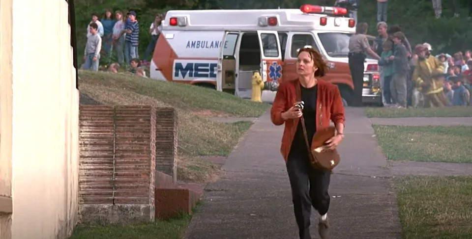 Person running with urgency near an ambulance and a crowd in the background, holding a camera, suggesting a dramatic or emergency scene