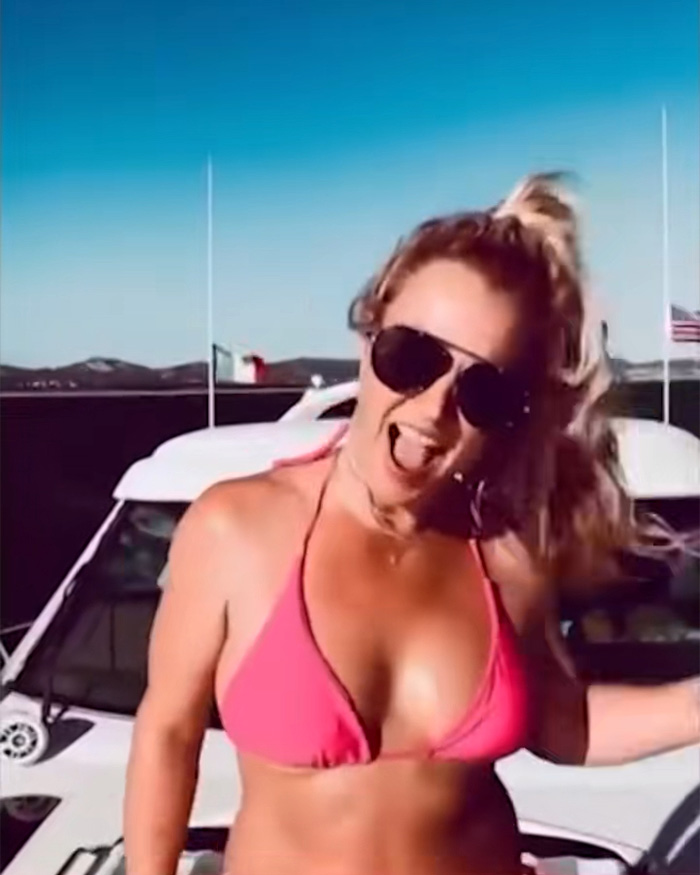 Britney Spears in pink bikini on a yacht wearing sunglasses, enjoying a sunny day with ocean and distant hills in the background.