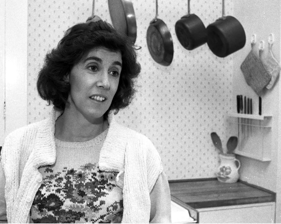 A person in a kitchen with hanging pots, wearing a patterned sweater and open cardigan, looking to the side