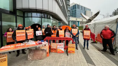BBC A group of people standing outside a glass building holding orange cards. They say things like "patients need doctors, doctors need jobs" and "doctors need jobs now". They are wearing orange bucket hats and beanies, and have an orange table with more signs leaning against it.