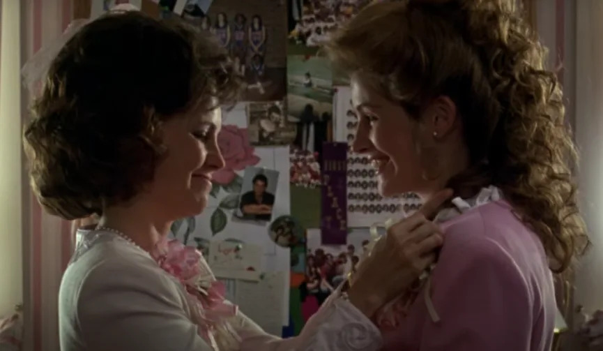 Two women smiling at each other, adjusting a corsage. They are in a room with a collage of photos on the wall behind them