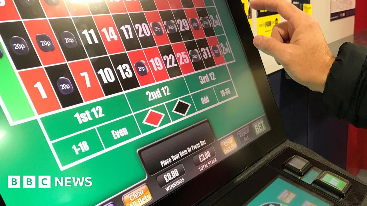 A man's hand is pictured about to press the brightly lit screen of a slot machine. The screen shows a series of red and black squares, some of which are filled with the price of bets placed. Details of stakes and winnings are also pictured.