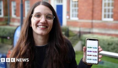 A woman with long brown hair, glasses and a dark jumper smiles as she holds up a phone with her left hand.