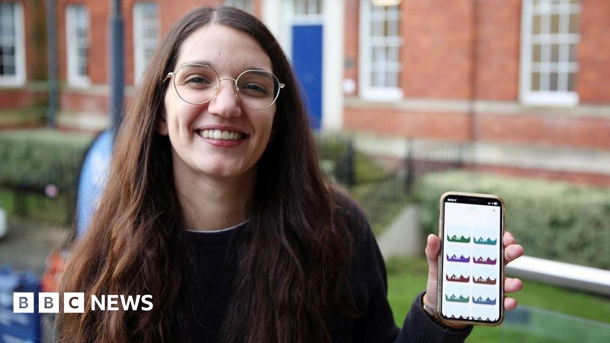 A woman with long brown hair, glasses and a dark jumper smiles as she holds up a phone with her left hand.