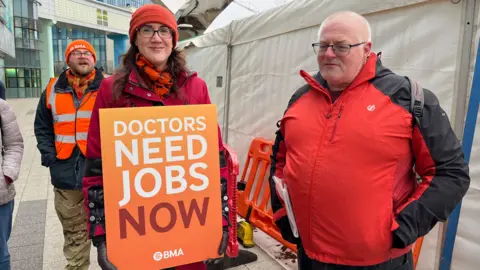 A woman with brown hair is wearing an orange beanie with a red coat and orange scarf. She is holding an orange card that reads "doctors need jobs now". There is a man with very short white hair standing next to her, and he is wearing a black and red coat