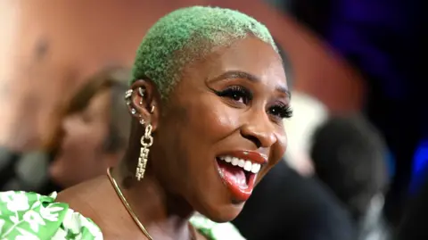 Getty Images Cynthia Erivo at the Washington DC premiere of Harriet in 2019. She is wearing a green and white print dress and her cropped hair is dyed green.