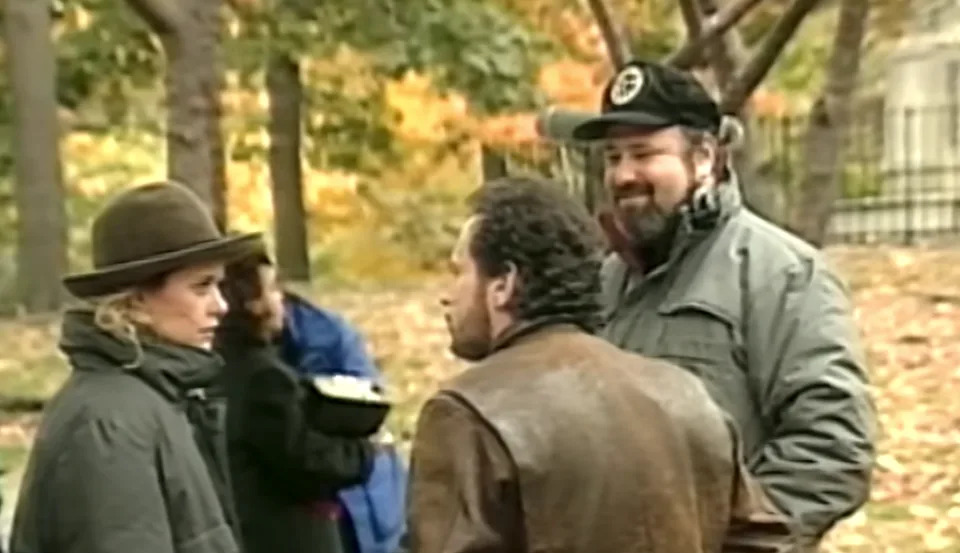 Three people in a park having a conversation, dressed warmly in coats and hats. Background shows autumn trees and a blurry person holding a bowl