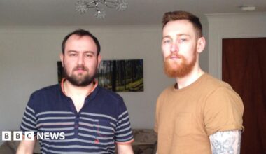 Two men stood in a living room. The man on the left has black hair, black beard, navy polo t-shirt with orange trim and grey stripes. The man on the right has red hair, a red beard, a light brown t-shirt and a heavily tattooed left arm. The living room is cream, with a cream settee, woodland photograph, dark wooden door and a flowery light fitting.