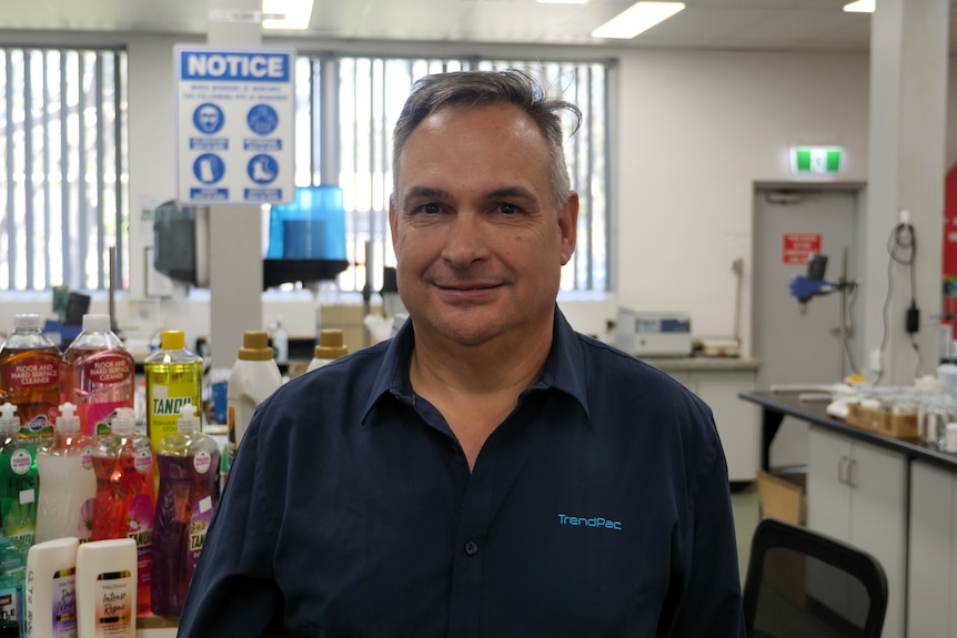 A man in a dark blue collared shirt. There are plastic bottles on a shelf behind him.