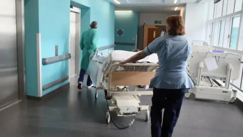 Getty Images Two hospital staff in uniform pushing an empty bed along a corridor