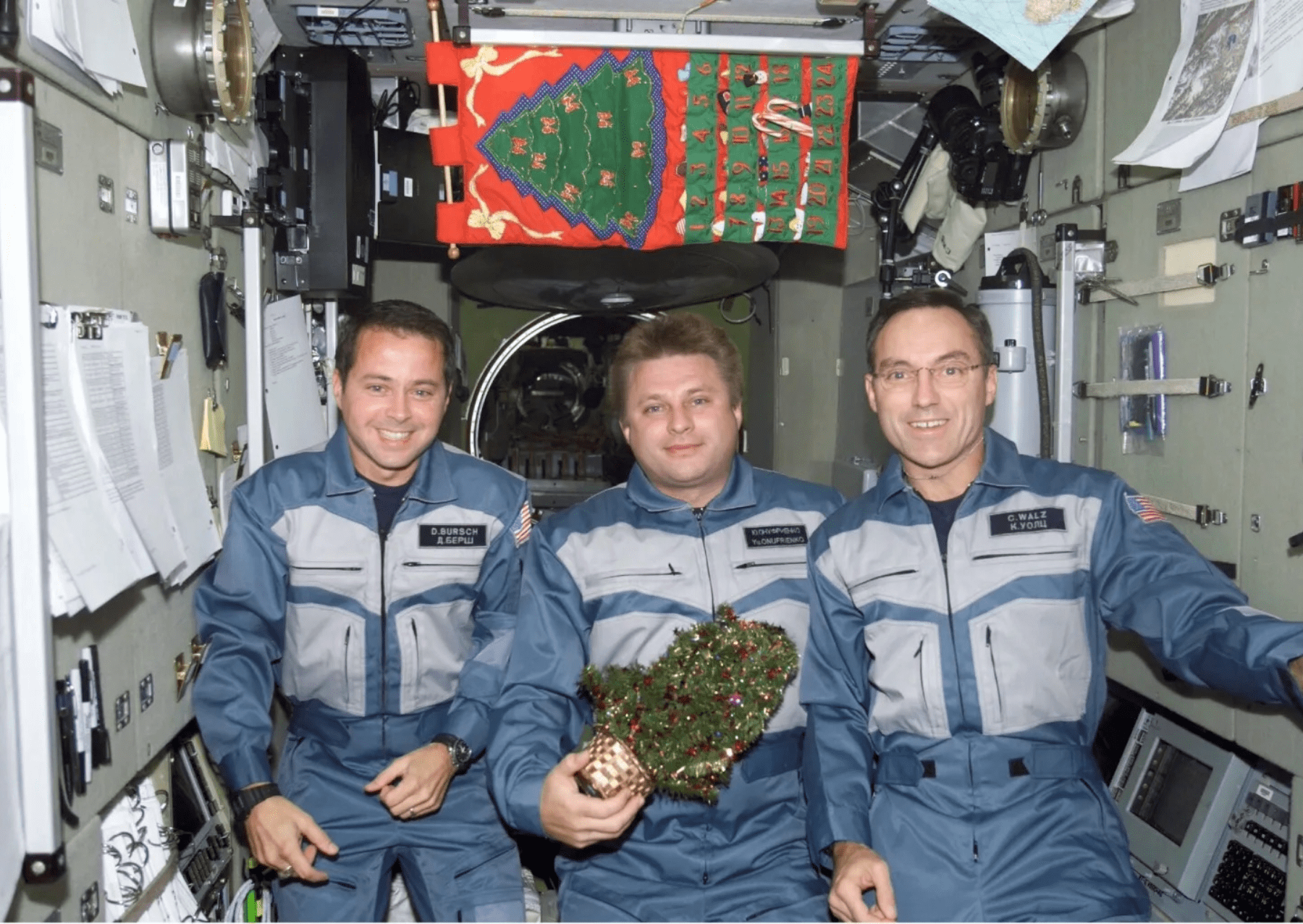 three male astronauts aboard the ISS, one holds a small christmas treee