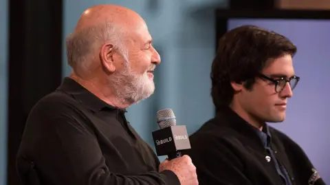 Getty Images Rob Reiner, wearing black shirt, in profile holding a microphone next to Nick Reiner, in dark jacket and wearing dark rimmed glasses, also in profile