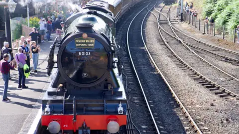 BBC A black steam train pulls into a railway station with photographers on the platform as it approaches