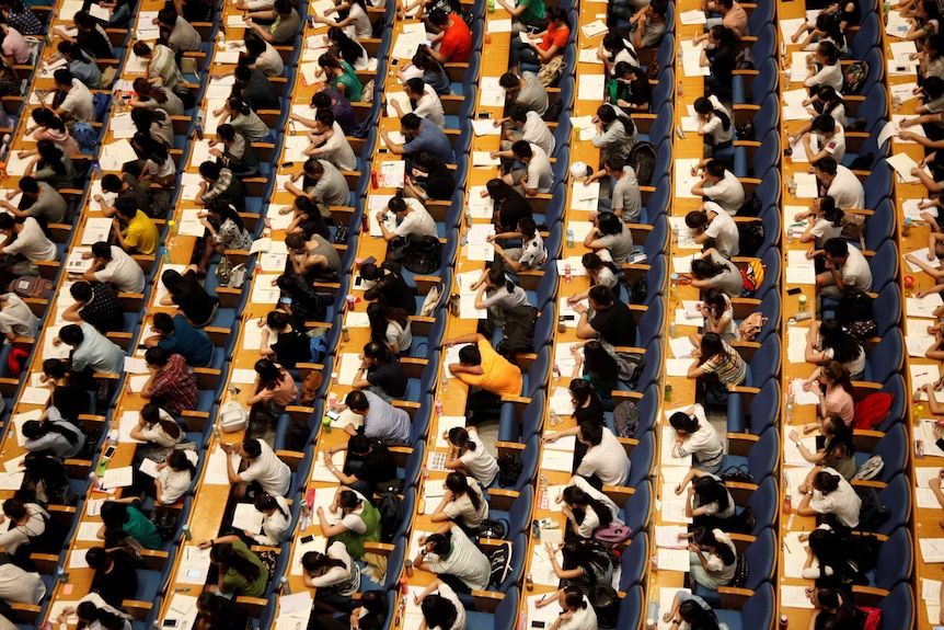 An overhead view of many rows of students sitting at bench desks.