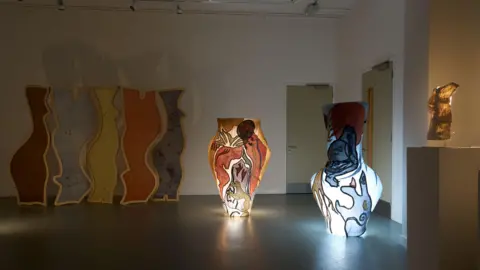 BETHLEM GALLERY The exhibition room which is fairly dark, there is a wooden screen in the back with arching shapes and different colours, there are two light sculptures on the side, one has a red tint and the other has a blue tint.