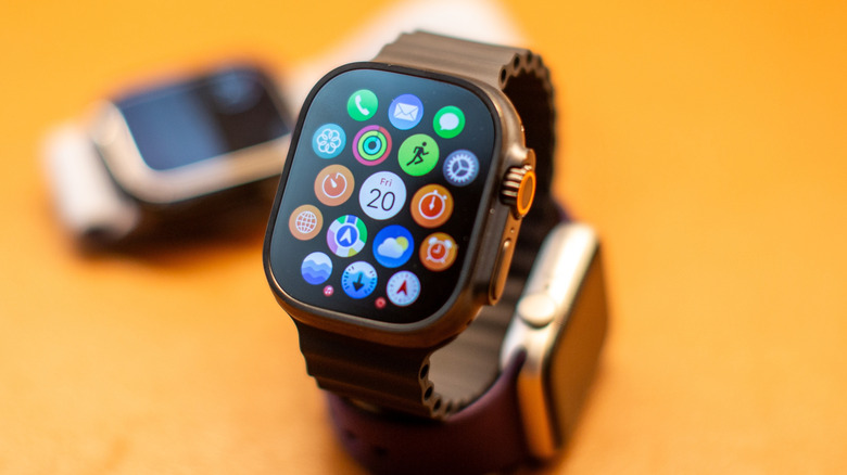 A closeup of an Apple Watch sitting upright on its wristband, two other Apple Watches can be faintly seen in the background.